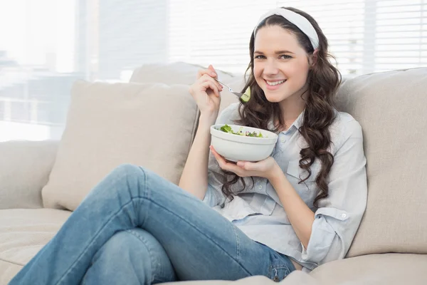 Довольно непринужденная женщина на уютном диване ест салат — стоковое фото