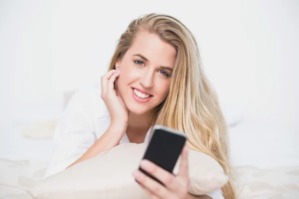 Uśmiechający się modelu gospodarstwa jej smartphone, leżąc na łóżku przytulne — Zdjęcie stockowe