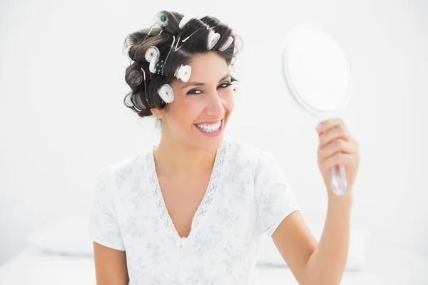 Счастливая брюнетка в роликах для волос с ручным зеркалом, улыбающаяся в камеру — стоковое фото