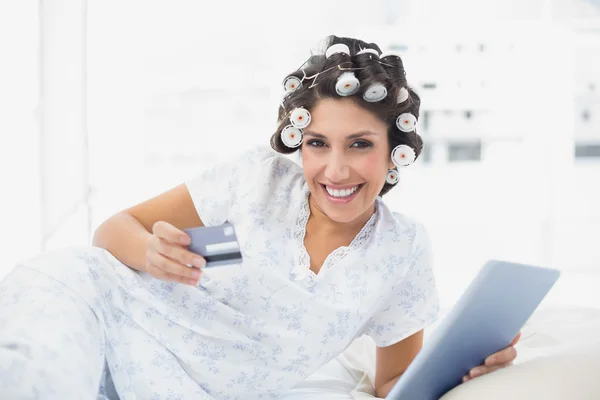 Улыбающаяся брюнетка в бигуди для волос лежит на кровати, используя планшет, чтобы ходить по магазинам онлайн — стоковое фото