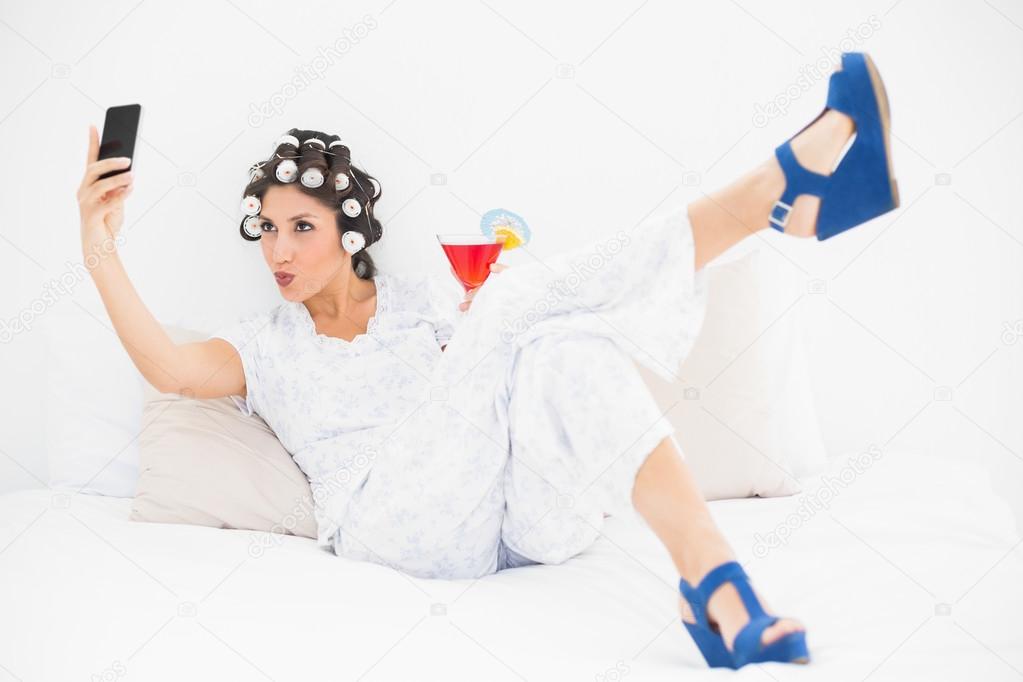 Brunette in hair rollers and wedge shoes having a cocktail taking a selfi