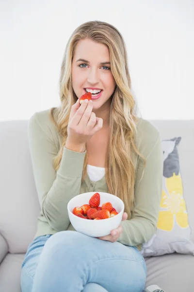 Vrolijke mooie blonde eten aardbeien zittend op de gezellige sofa — Stockfoto