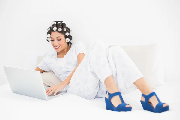 Улыбающаяся брюнетка в роликах для волос и клиновых туфлях с помощью ноутбука на кровати — стоковое фото