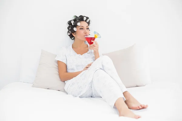 Smiling brunette in hair curlers sitting on her bed drinking a cocktail — Stock Photo, Image