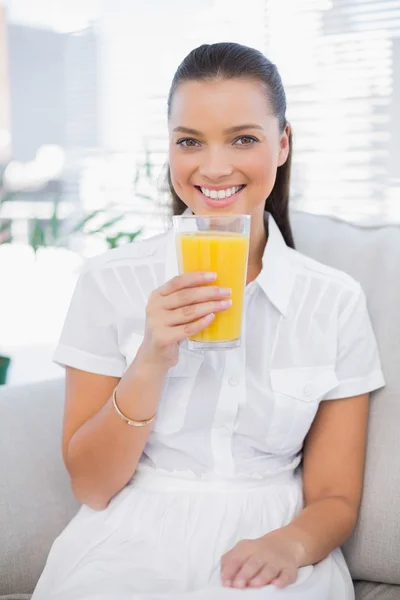 Mujer bonita sonriente sosteniendo jugo de naranja sentado en un sofá acogedor —  Fotos de Stock