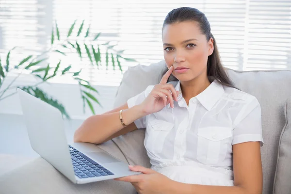 Mujer bonita reflexiva usando el ordenador portátil sentado en el sofá acogedor — Foto de Stock