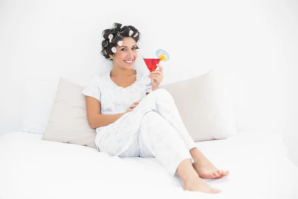 Smiling brunette in hair curlers sitting on her bed holding a cocktail — Stock Photo, Image