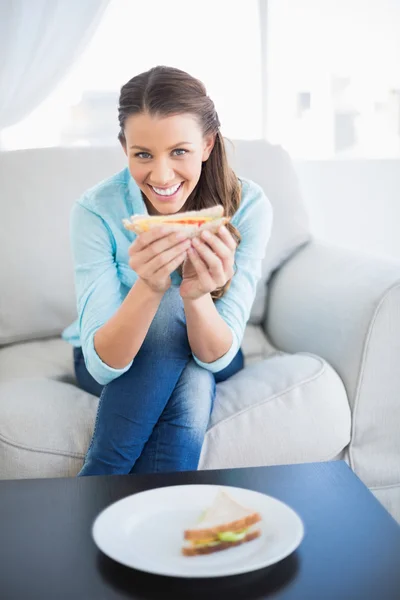 Happy woman sitting on sofa showing sandwich at camera — Stock Photo, Image