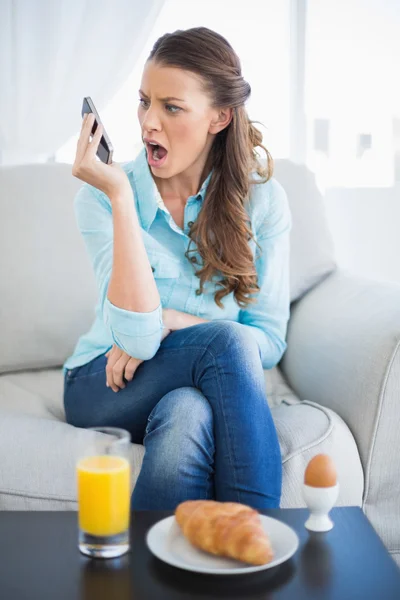 Mujer enojada gritando en el teléfono sentado en el sofá —  Fotos de Stock