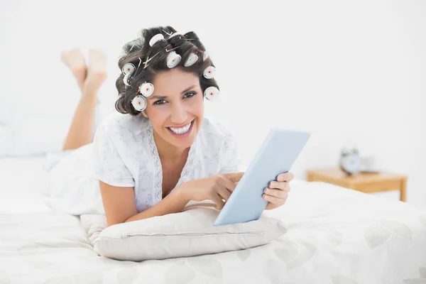 Lächelnde Brünette in Haarrollen, die mit ihrem Tablet auf ihrem Bett liegt — Stockfoto
