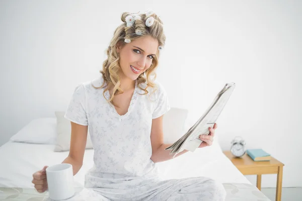 Smiling cute blonde wearing hair curlers holding newspaper — Stock Photo, Image