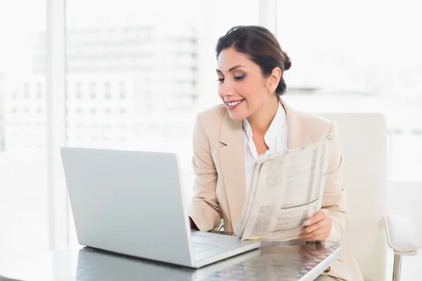 Cheerful businesswoman holding newspaper while working on laptop — Stock Photo, Image