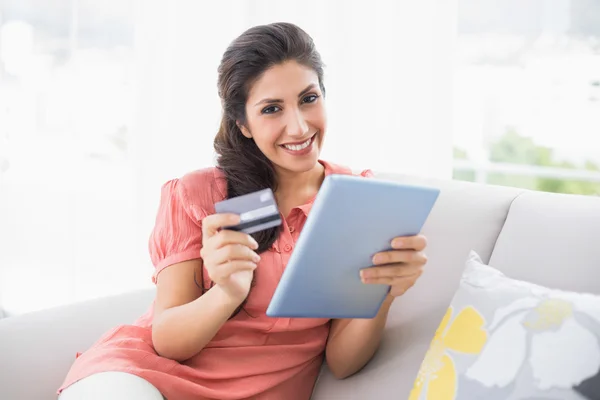 Lächelnde Brünette, die auf ihrem Sofa sitzt und mit dem Tablet online einkauft — Stockfoto