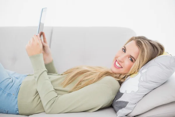 Joyeux jolie blonde en utilisant sa tablette couchée sur un canapé confortable — Photo