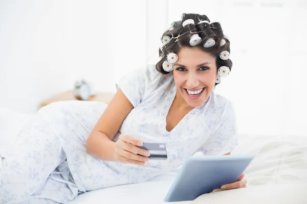 Happy brunette in hair rollers lying on her bed using her tablet to shop online — Stock Photo, Image