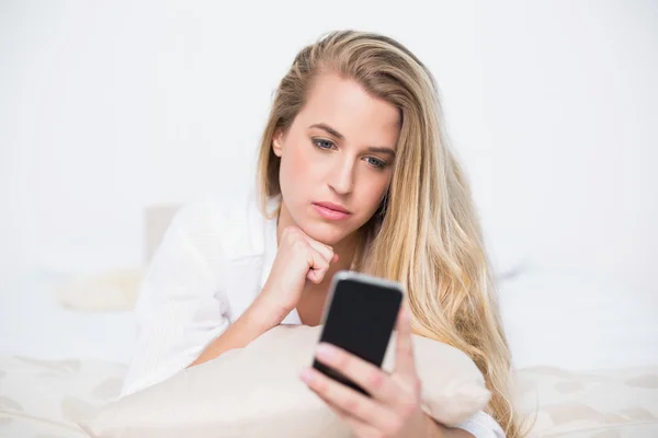 Nachdenkliches Traummodel blickt auf ihr Smartphone, das auf kuscheligem Bett liegt — Stockfoto
