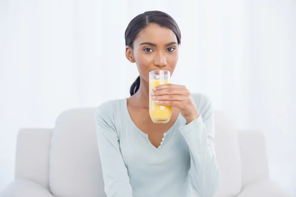 Mulher sorridente sentada no sofá acolhedor bebendo suco de laranja — Fotografia de Stock