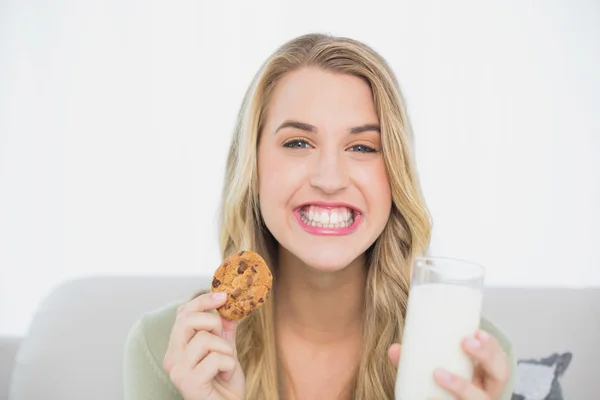 Happy cute blonde eating cookie with milk sitting on cosy sofa — Stock Photo, Image