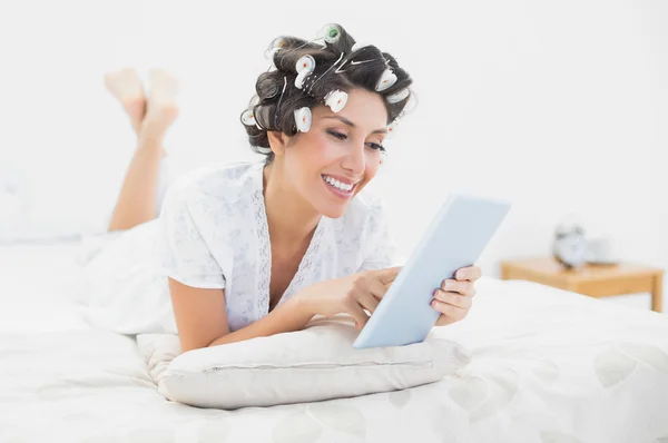 Morena feliz en rodillos de pelo acostada en su cama usando su tableta —  Fotos de Stock