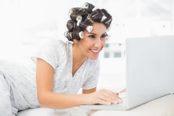 Smiling brunette in hair rollers lying on her bed using her laptop — Stock Photo, Image