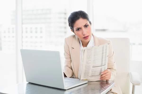 Achtersteven zakenvrouw krant bedrijf terwijl u werkt aan laptop camera kijken — Stockfoto