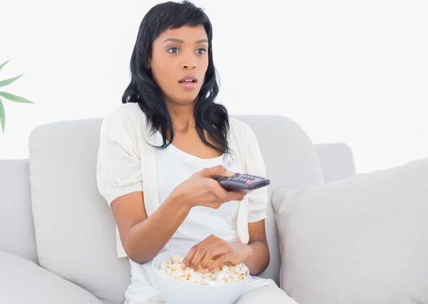 Shocked black haired woman in white clothes watching tv while eating popcorn — Stock Photo, Image