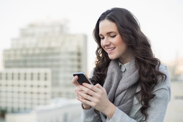 Glad söt brunett i vinterkläder skicka en text på sin smartphone — Stockfoto