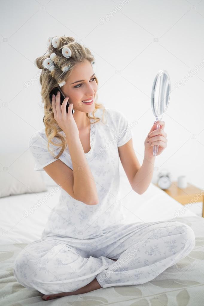 Happy blonde wearing hair curlers looking at reflection while ph