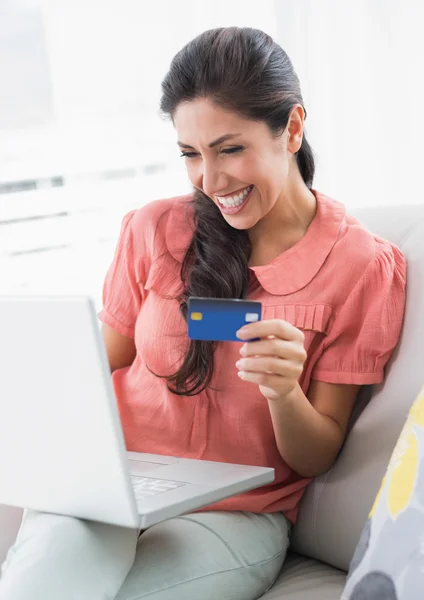 Fröhliche Brünette sitzt auf ihrem Sofa mit Laptop, um online einzukaufen — Stockfoto