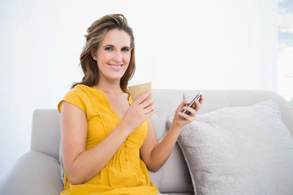Mujer sonriente sentada en el sofá holing café y el uso de teléfono — Foto de Stock