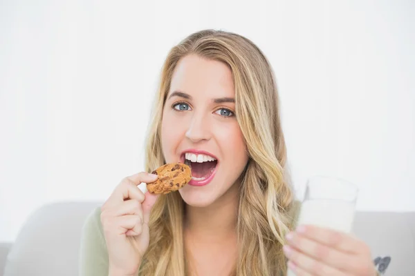 Veselá roztomilá blondýnka jíst cookie s mlékem na pohodlná pohovka — Stock fotografie