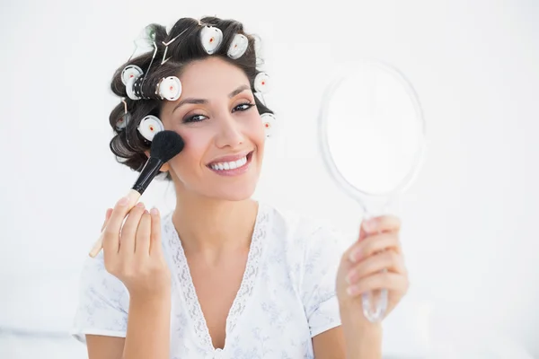 Sonriente morena en rodillos de pelo sosteniendo espejo de mano y aplicando maquillaje —  Fotos de Stock