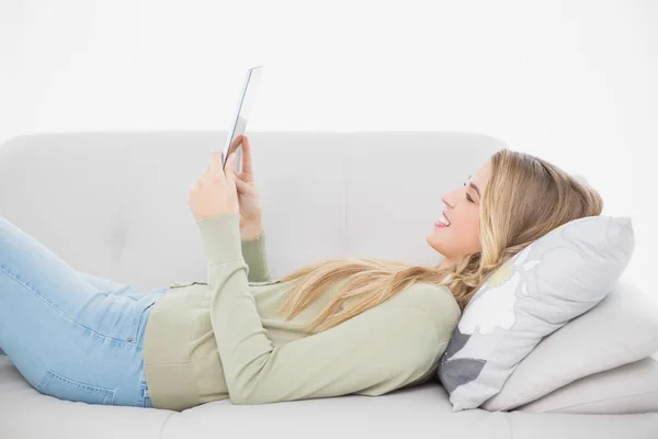 Smiling pretty blonde using her tablet lying on cosy sofa — Stock Photo, Image