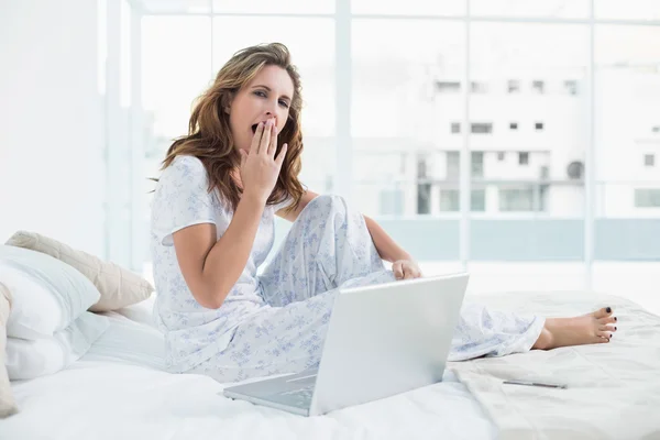 Trött kvinna som sitter på mysiga säng med laptop — Stockfoto