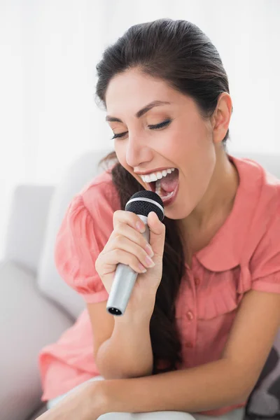 Brunette sitting on her sofa singing into microphone — Stock Photo, Image