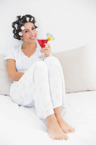 Веселая брюнетка в бигуди, сидящая на кровати, держа коктейль — стоковое фото