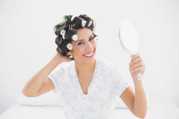 Smiling brunette in hair rollers holding hand mirror — Stock Photo, Image