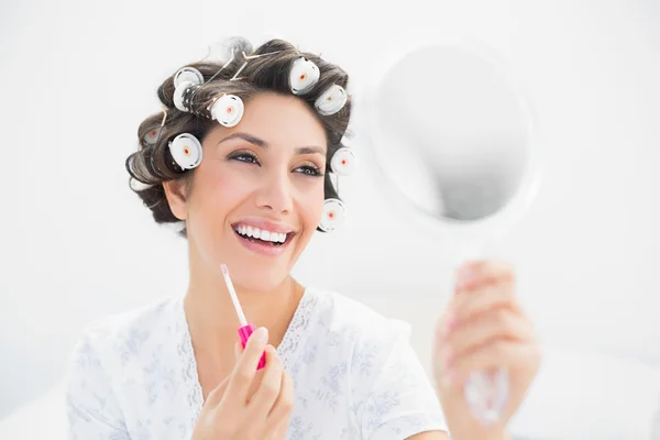Happy brunette in hair rollers looking in hand mirror and holding lip gloss — Stock Photo, Image