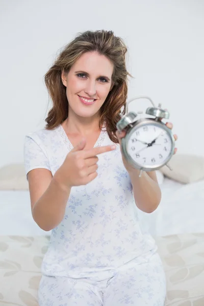 Lachende vrouw die zit op bed wijzend op alarm clock — Stockfoto