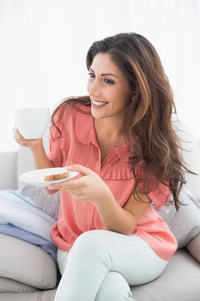 Pretty brunette sitting on her sofa holding cup and saucer — Stock Photo, Image