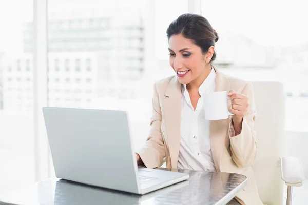 Alegre mujer de negocios sosteniendo taza mientras trabaja en el ordenador portátil —  Fotos de Stock