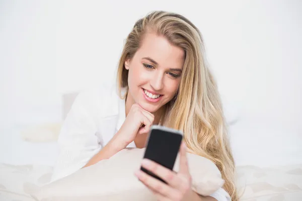 Sorrindo modelo lindo olhando para seu smartphone deitado na cama acolhedora — Fotografia de Stock