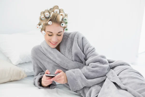 Happy relaxed blonde woman in hair curlers using her mobile phone — Stock Photo, Image