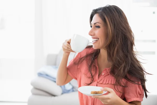 Glückliche Brünette sitzt auf ihrem Sofa und hält Tasse und Untertasse mit einem Keks — Stockfoto