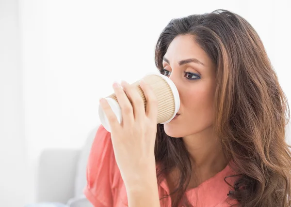 Smiling brunette sitting on her sofa sipping from disposable cup — Stock Photo, Image