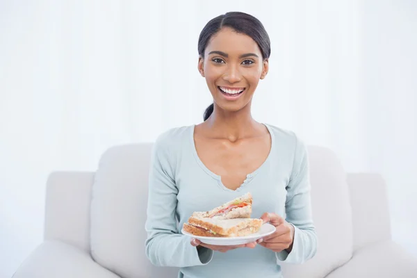 Mujer atractiva sonriente sentada en un acogedor sofá sosteniendo un sándwich — Foto de Stock