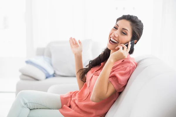 Riendo morena sentada en su sofá en el teléfono —  Fotos de Stock