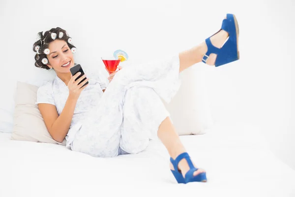 Brunette in hair rollers and wedge shoes having a cocktail sending a text on bed — Stock Photo, Image