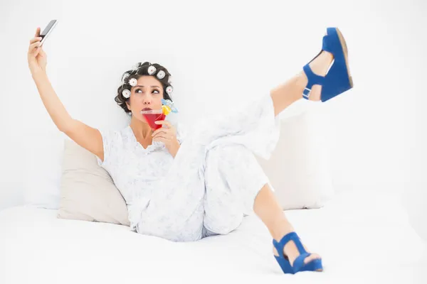 Brunette in hair rollers and wedge shoes drinking a cocktail taking a selfi — Stock Photo, Image