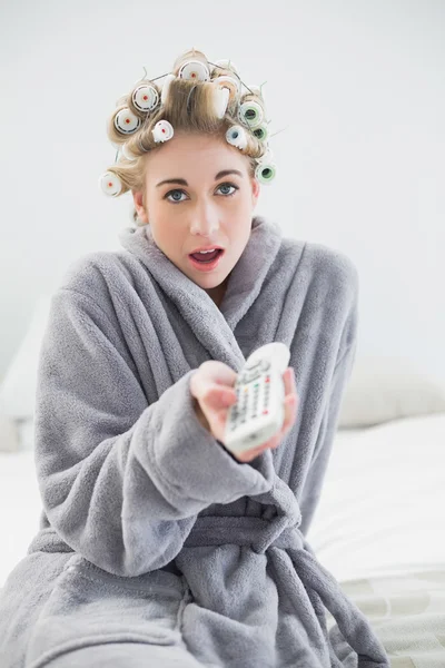 Surprised relaxed blonde woman in hair curlers using a remote control — Stock Photo, Image